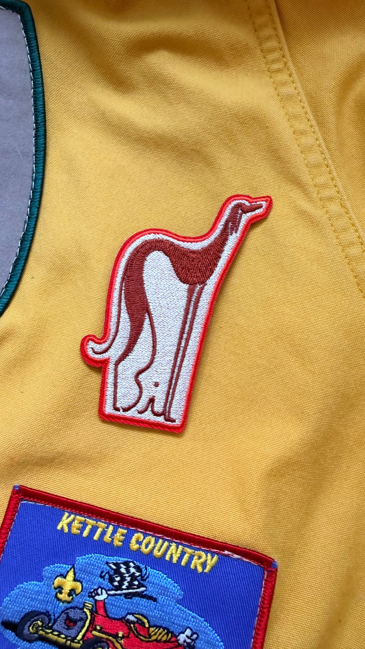 Close-up of a mustard yellow garment featuring an embroidered kangaroo patch with a red border and a blue 'KETTLE COUNTRY' patch below it.