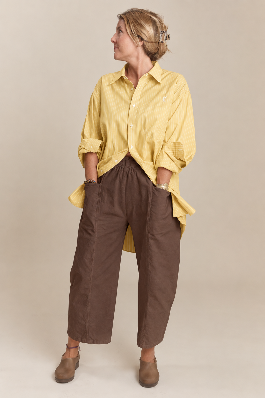 A woman modeling a yellow button-up shirt and brown wide-leg cropped pants against a plain beige studio background.