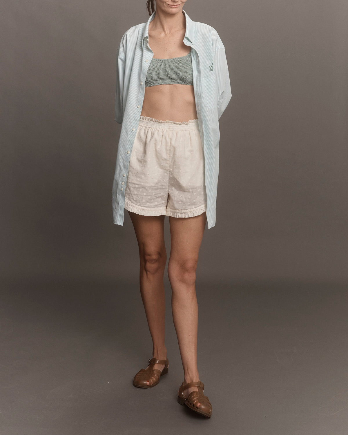 Woman wearing a light blue shirt, green sports bra, beige shorts, and brown sandals on a gray background