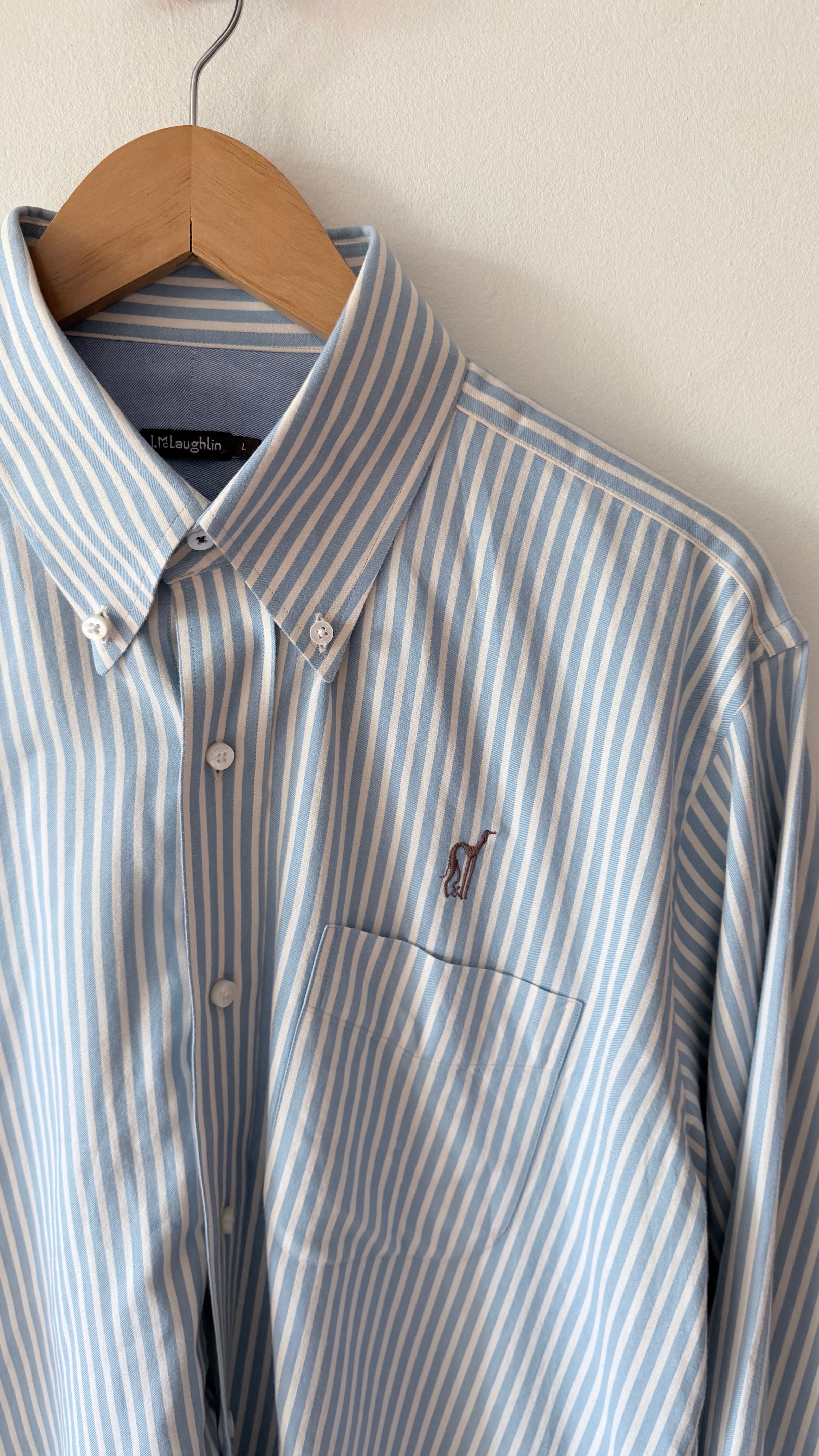 A light blue and white vertically striped button-down shirt by Ralph Lauren, featuring a small embroidered polo player logo on the chest, hanging on a wooden hanger against a plain off-white wall.