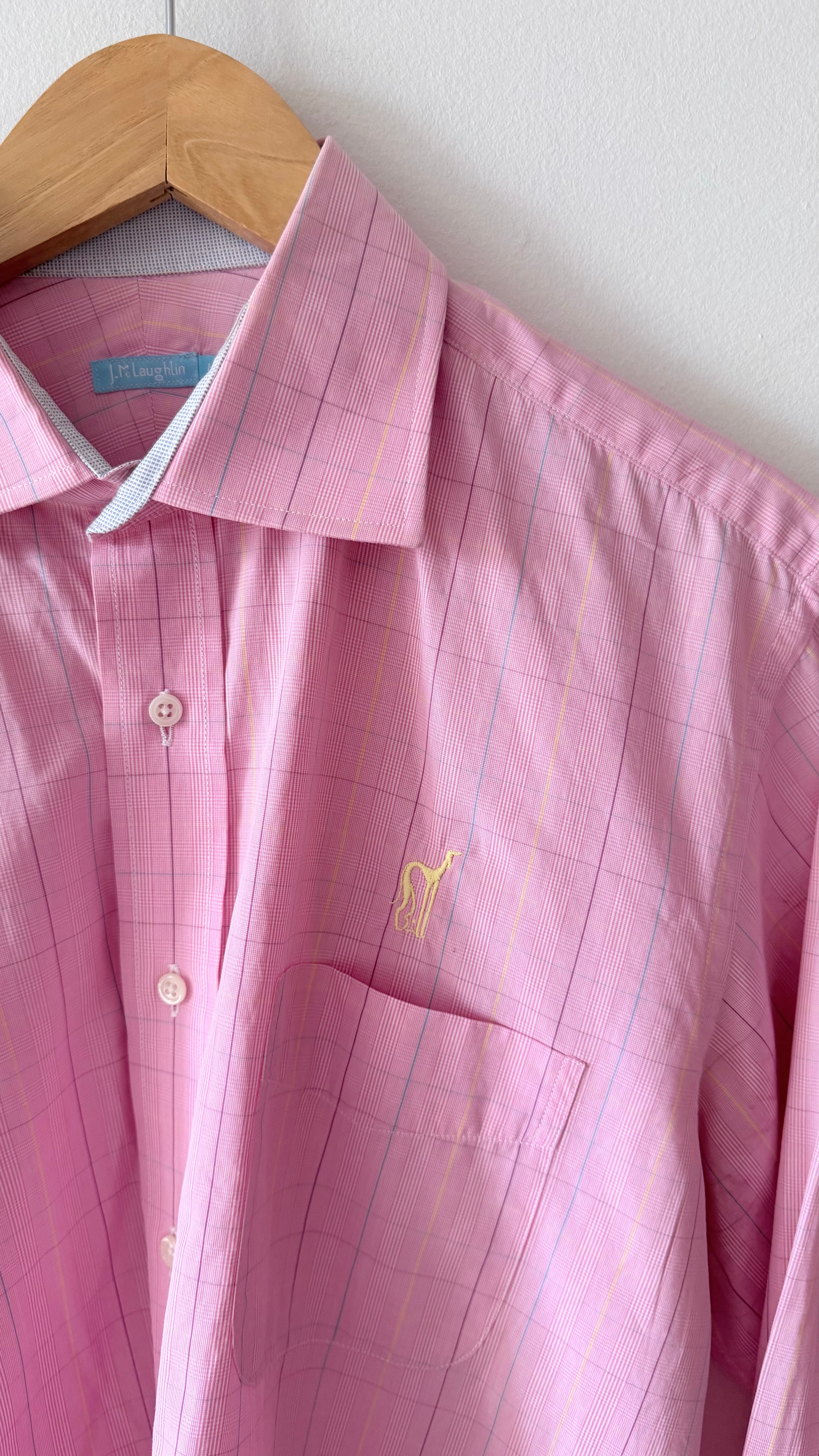 A pink long-sleeve button-up shirt with a subtle windowpane grid pattern, hanging on a wooden hanger against a white wall. It features a single chest pocket with a small yellow embroidered polo player logo, indicating the brand Polo Ralph Lauren.