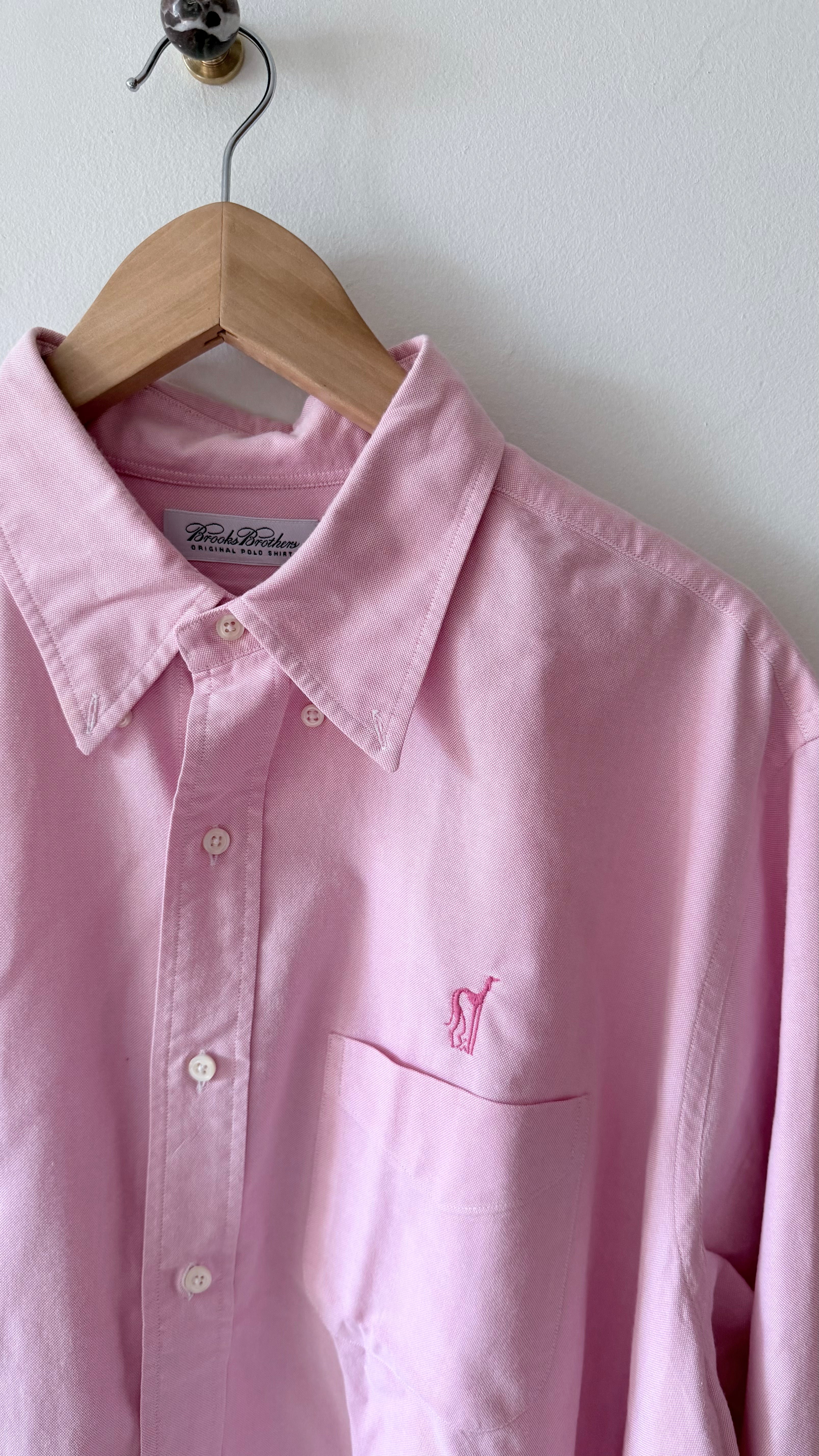 A light pink long-sleeve button-up shirt with a small embroidered polo player logo on the chest pocket, hanging on a wooden hanger against a white wall.