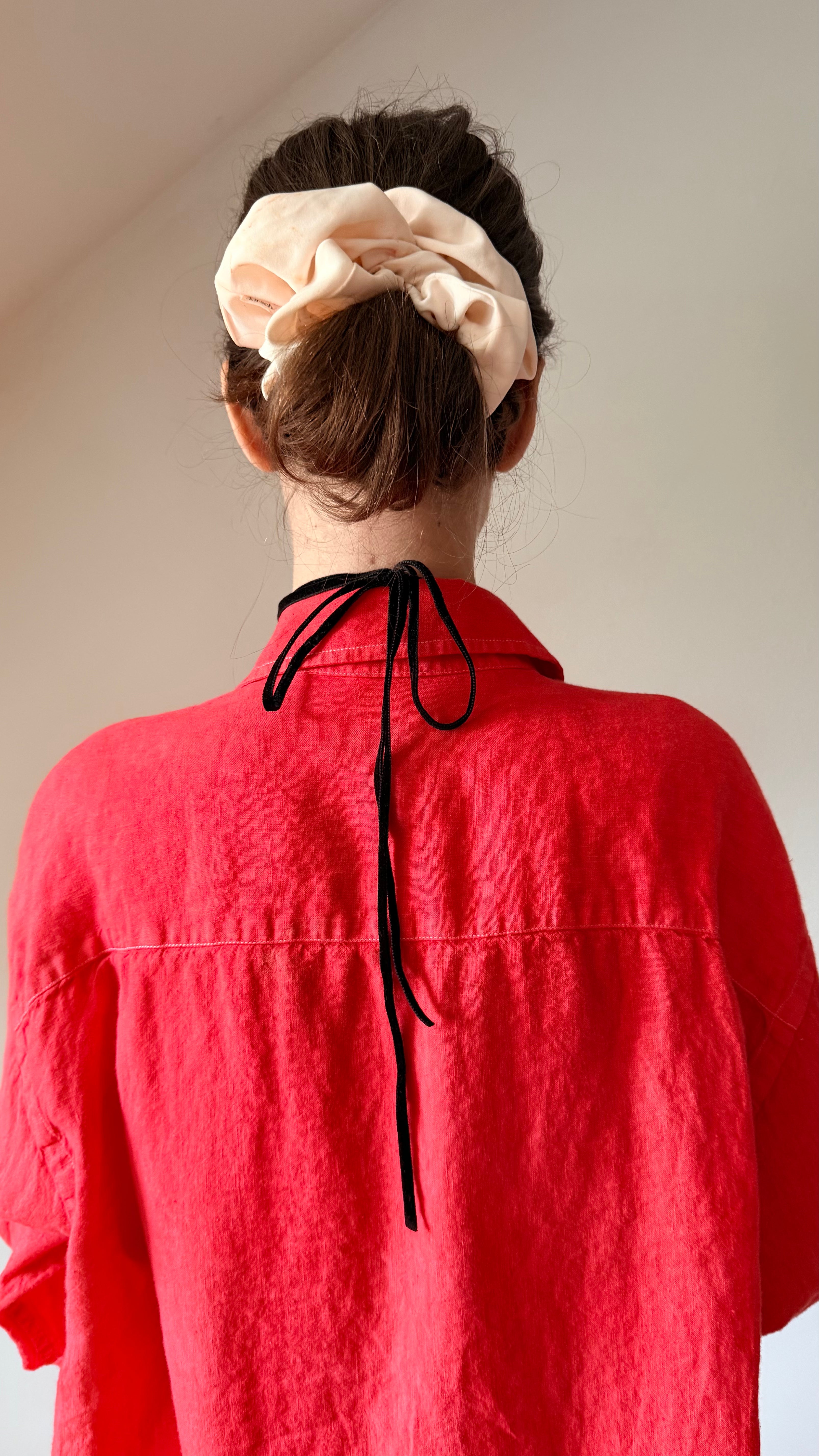 A person shown from behind wearing a bright red linen shirt and a cream-colored fabric scrunchie, photographed against a plain off-white wall.