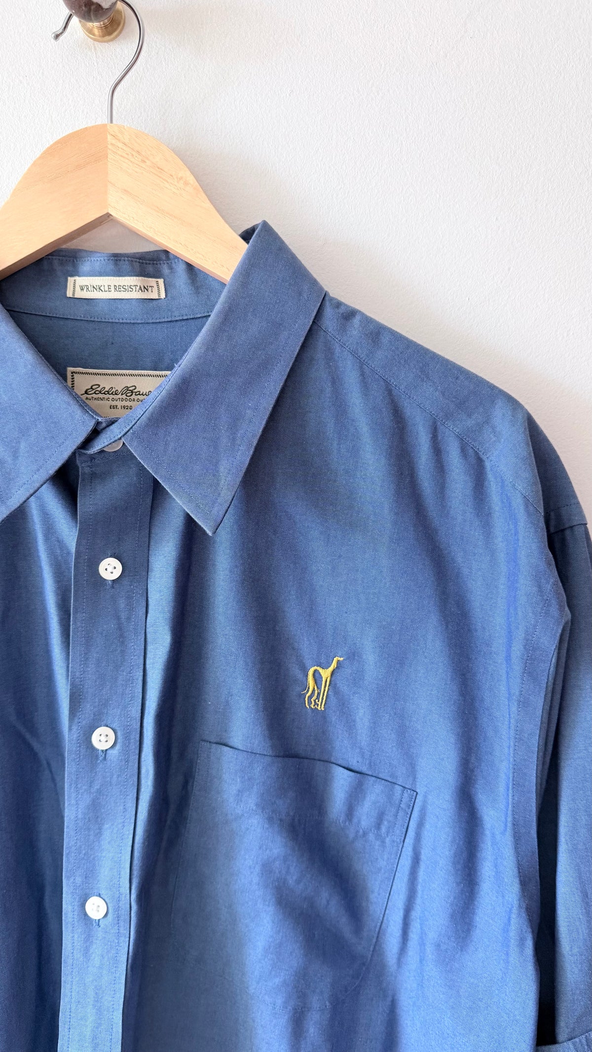 A blue long-sleeve button-up shirt hanging on a wooden hanger against a plain white wall. The shirt features a small yellow embroidered polo player logo on the left chest, with a visible Ralph Lauren neck label.