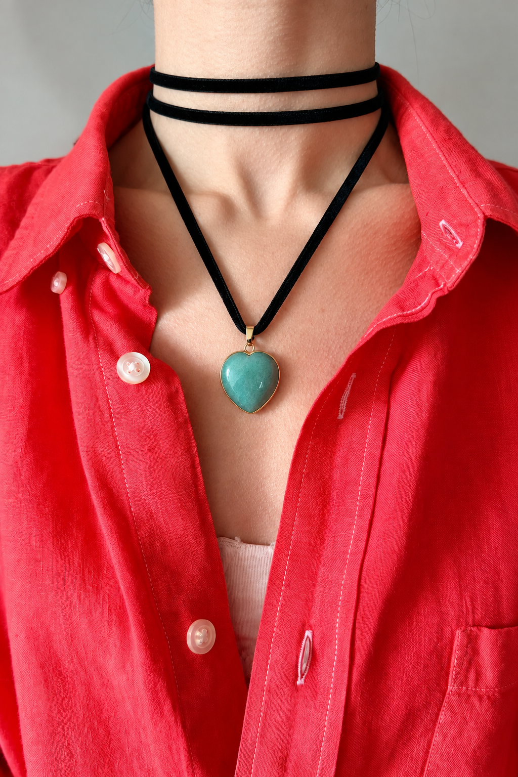 A close-up of a person wearing a black cord choker necklace with a heart-shaped turquoise pendant, layered over a red button-up shirt.